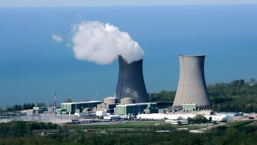 Nuclear power plant cooling towers beside a body of water with steam rising from one tower under a blue sky